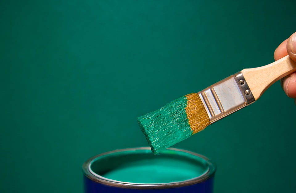 Un barattolo di vernice verde aperto con sopra un pennello intinto nello smalto e stretto da una mano, il tutto su sfondo dello stesso verde