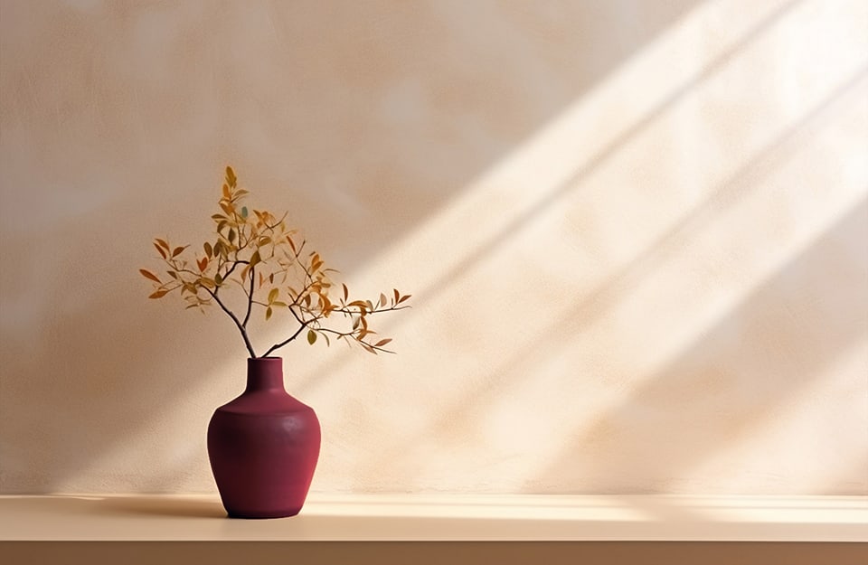 Parete color neutro-beige con davanti un vaso in ceramica color bordeaux che contiene un rametto decorativo con foglie ingiallite
