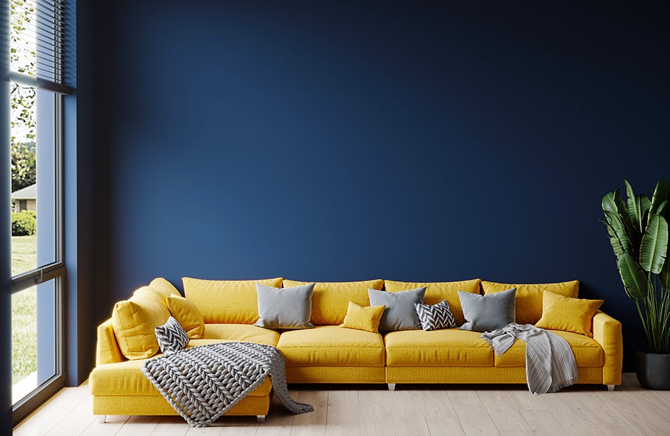 Un grande divano d'angolo giallo dalle linee squadrate, con cuscini grigi e coperte grigie, accanto a una grande finestra, in una stanza dalle pareti blu cobalto, pavimento in parquet e una pianta in vaso accanto al sofà