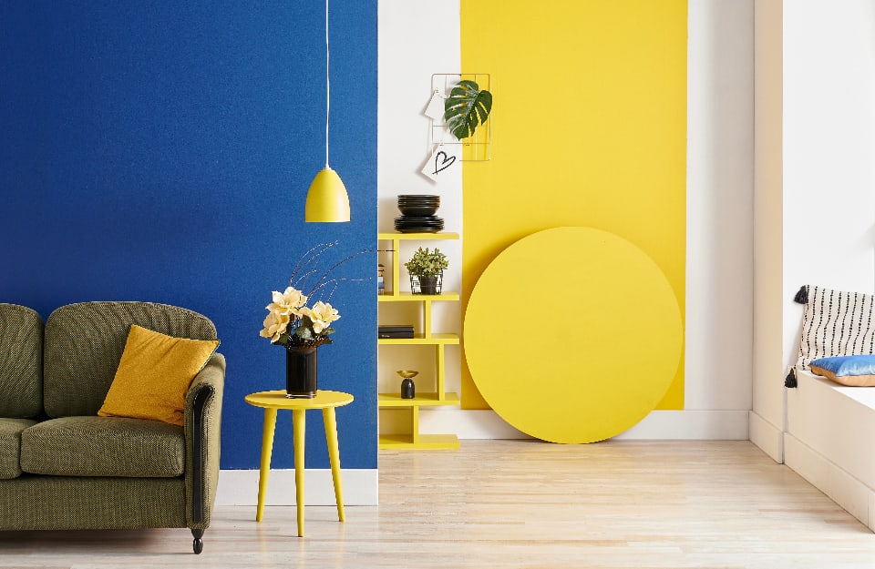 Angolo di una casa moderna, con parete blu, sofà verde, tavolino giallo e lampada gialla. Si intravede, dietro, un altro ambiente, con pareti bianche e gialle e una libreria gialla
