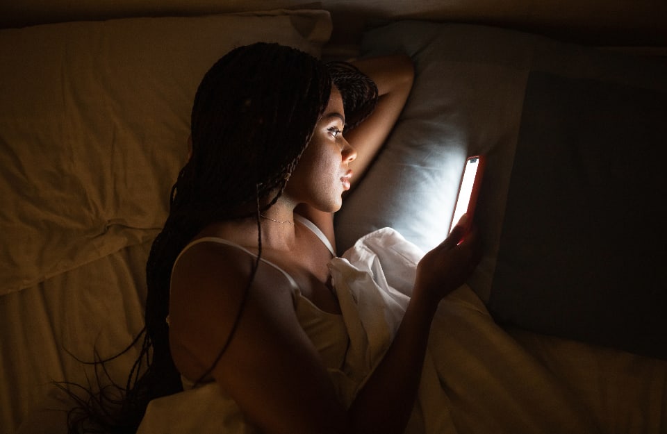 Una ragazza nera ha il viso illuminato dallo schermo dello smartphone che sta guardando mentre è sdraiata al letto sotto le coperte