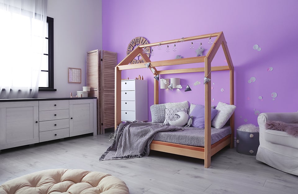 Camera da letto per bambino con una parete color glicine e struttura del letto in legno naturale a forma di casa