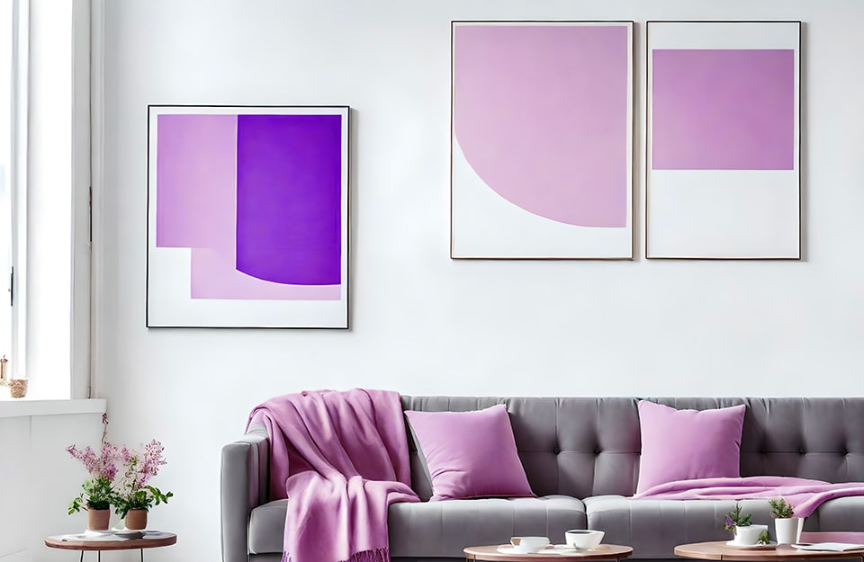 Dettaglio di un luminoso salotto moderno con pareti bianche, sofà trapuntato grigio con cuscini e coperte color glicine e quadri alle pareti con stampe a blocchi di colore geometrici sul glicine e sul viola
