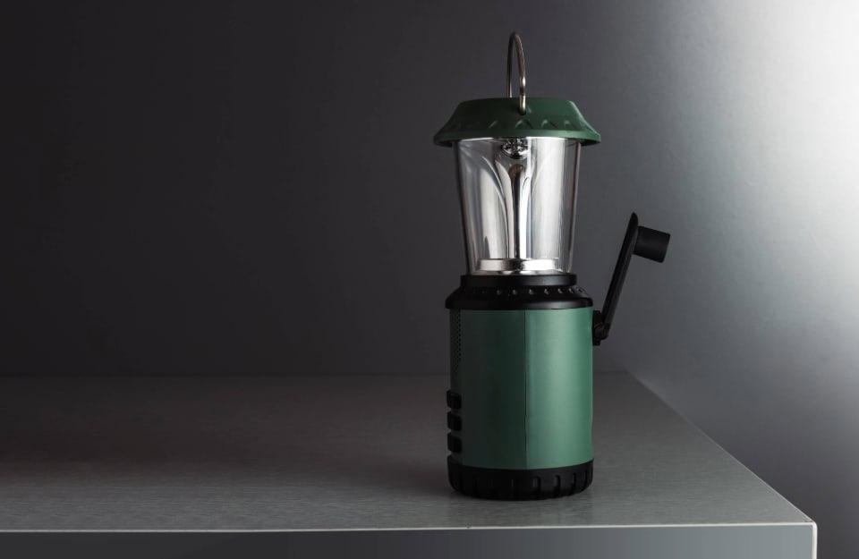 Una lampada da campeggio verde con luce a led e manovella per il caricamento manuale, sopra una lastra di cemento in uno sfondo monocromatico scuro