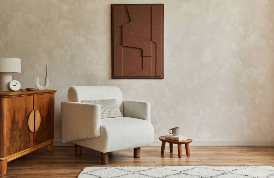 Angolo di un salotto moderno, con pareti beige a effetto limewash, poltrona dalla linee curve, quadro astratto materico, consolle vintage in legno