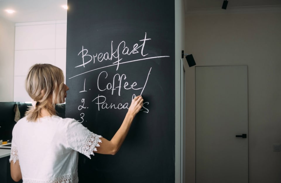 A woman is writing on a blackboard wall in her kitchen what is available for breakfast: coffee and pancakes