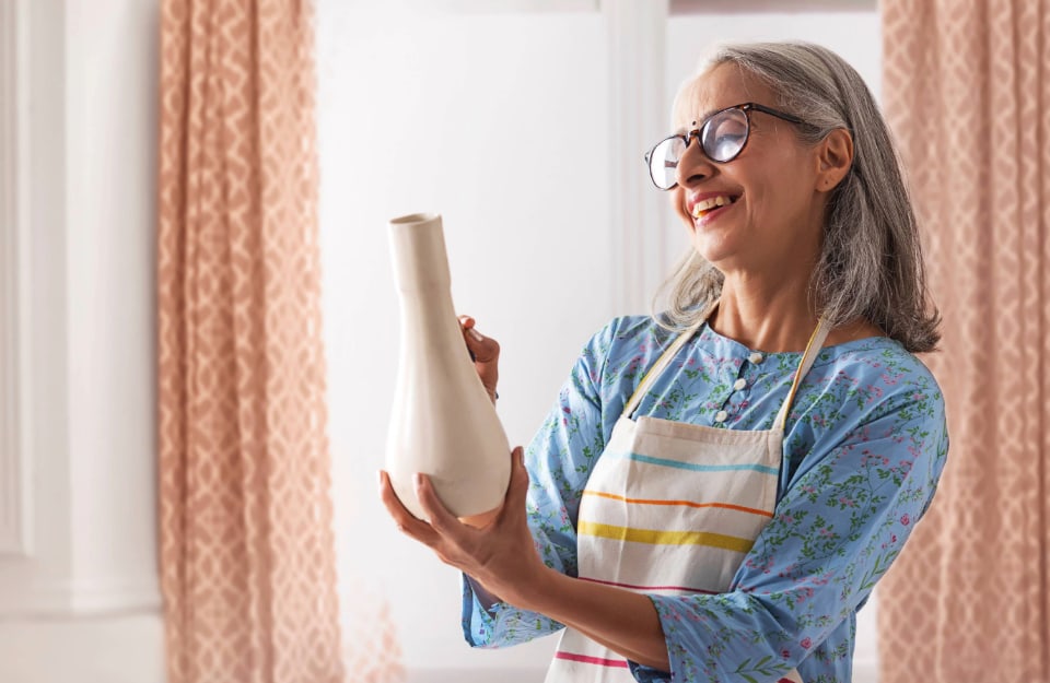 Una sorridente donna di mezza età con gli occhiali e il grembiule da lavoro sta dipingendo un lungo vaso bianco con un pennellino
