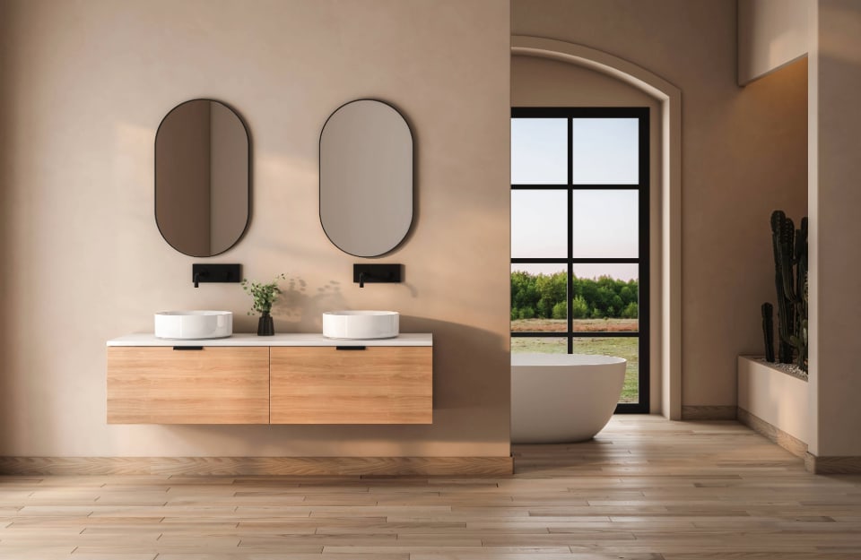 Una stanza da bagno luminosa e ampia, con pareti color écru, due specchi, mobili in legno e una vasca da bagno bianca che si intravede nell'ambiente attiguo, davanti a una grande vetrata