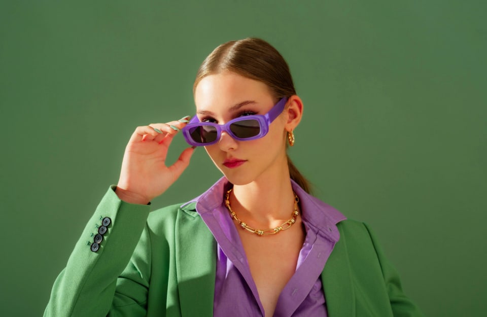 Una ragazza vestita con giacca verde e camicia viola, fa per togliersi gli occhiali viola, su sfondo monocromatico color verde