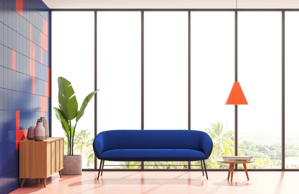 Luminoso salotto con grande finestra lungo tutta una parete, divano di design blu minimale, lampada con paralume rosso conico, consolle, in legno e un'altra parete rivestita di piastrelle blu, con piastrelle rosse a disegnare motivi tipo Tetris
