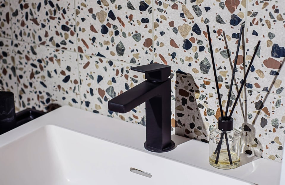Dettaglio di un bagno con lavandino in ceramica bianca e rubinetto nero dalle linee essenziali, con muro rivestito in piastrelle a effetto terrazzo alla veneziana