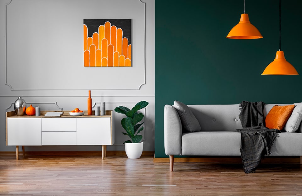 Salotto con parquet, parete bianca decorata con cornici, parete verniciata verde petrolio, sofà grigio, lampadari arancioni che richiamano il colore di un quadro astratto appeso alla parete sopra a una consolle bianca
