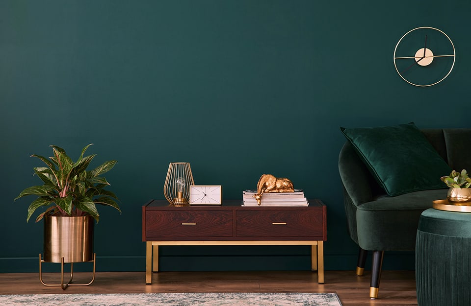 Dettaglio di un salotto con parete color verde petrolio, parte di un sofà dello stesso colore, tavolinetto con cassetti e struttura in metallo, orologio metallico art decó alla parete e vaso metallico con pianta