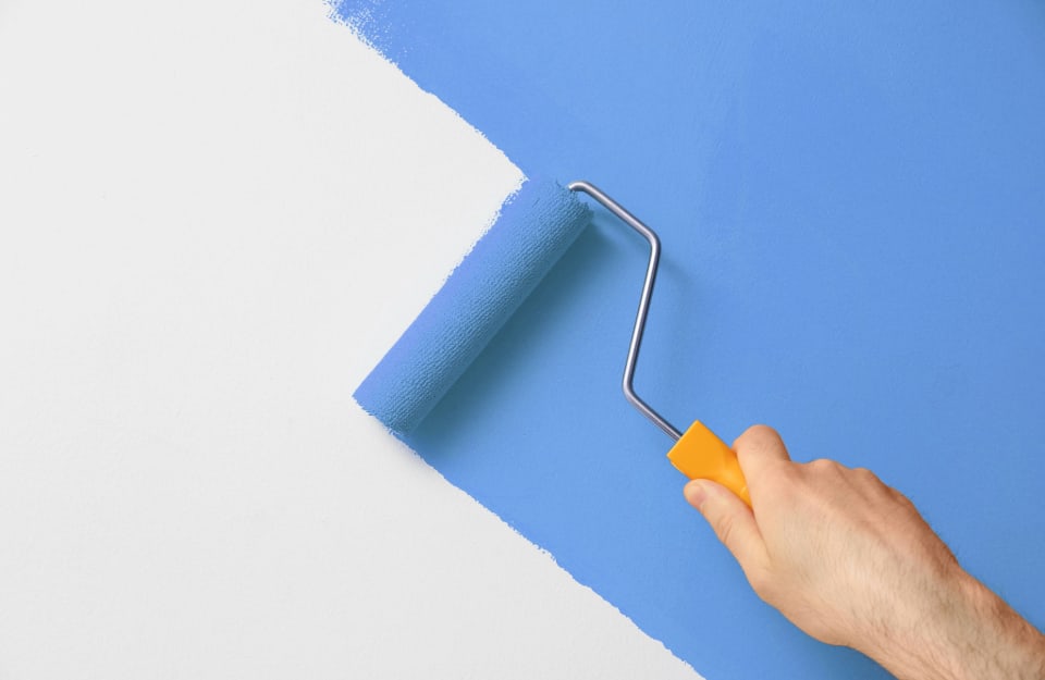 Una mano sta dipingendo di color blu fiordaliso, con un piccolo rullo, in diagonale, una parete bianca