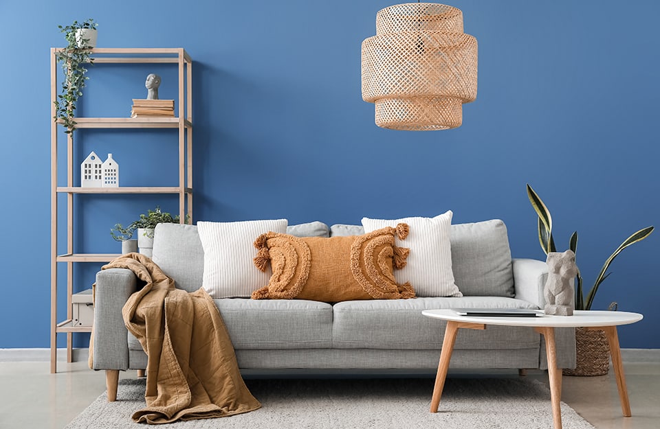 Scandinavian-style living room with cornflower blue wall, light grey sofa with wooden frame, white, grey and brown cushions, white round coffee table with natural wood legs, wicker chandelier, open bookcase in natural wood with books and knick-knacks