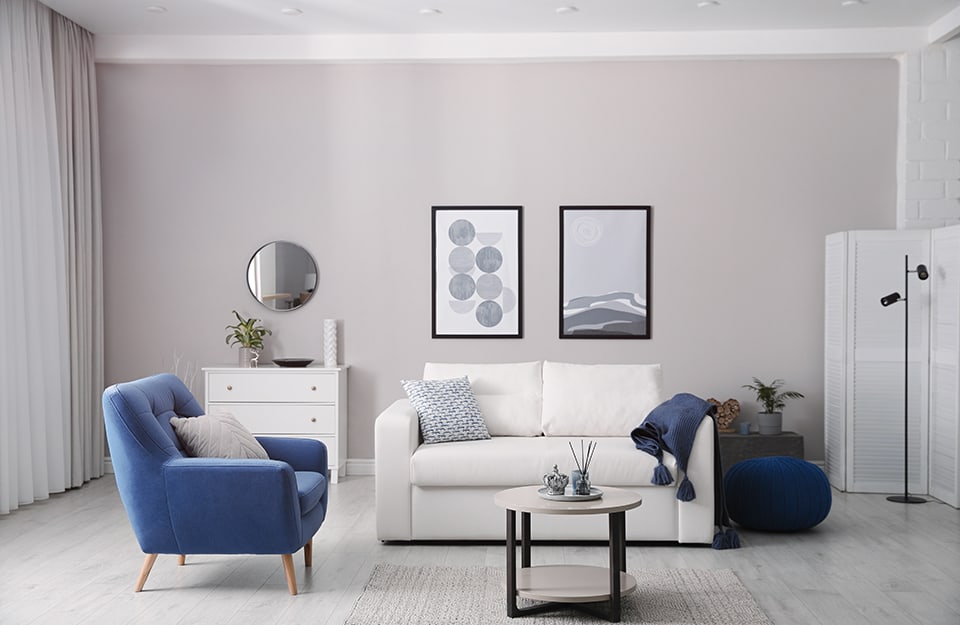 Sala de estar moderna y esencial con sofá, sillones, mesa de centro, lámpara de pie, tabique de madera, cómoda, alfombra, espejo y cuadros, todo en tonos blancos y grises, con detalles en negro, y un sillón azul aciano