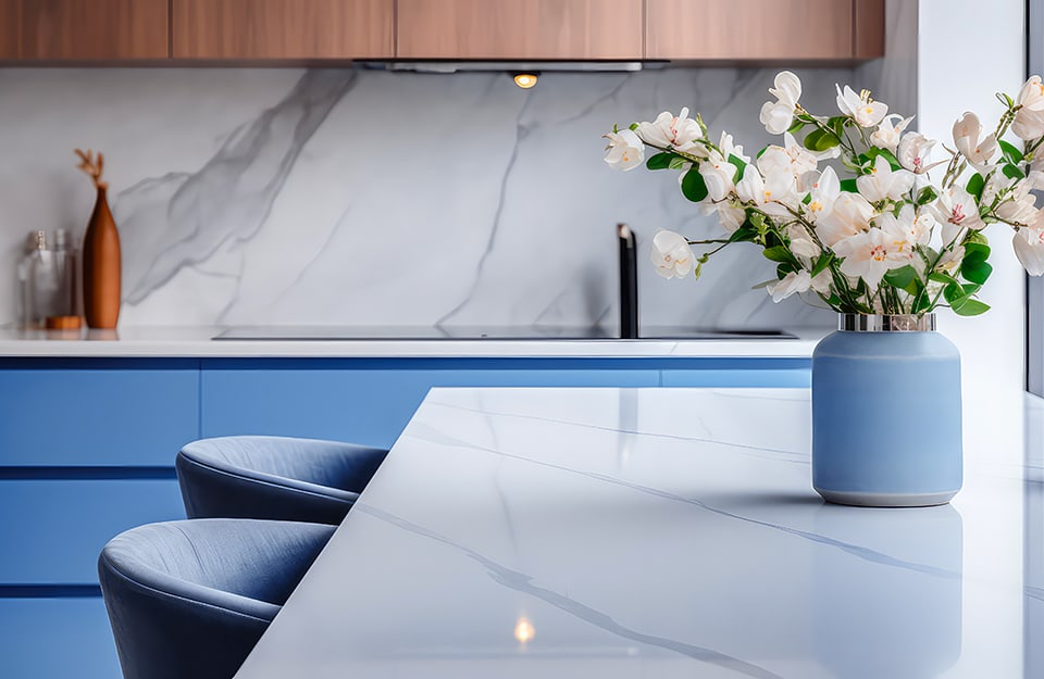 Dettaglio di una cucina sui colori del blu fiordaliso e del legno naturale, con isola con top in marmo bianco, sgabelli blu fiordaliso, vaso azzurro chiaro con rametti di fiori
