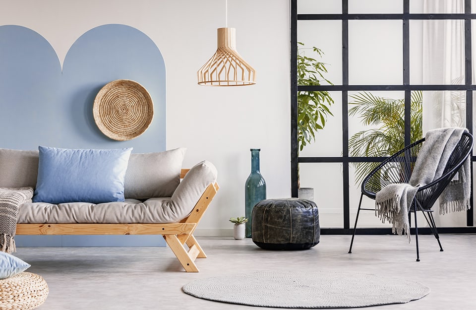Sala de estar moderna con gran ventanal con marco de rejilla negro, pared blanca con decoración de arcos azul aciano, sofá de madera de estilo escandinavo con cojín azul aciano, lámpara de mimbre, sillón y diversos objetos decorativos