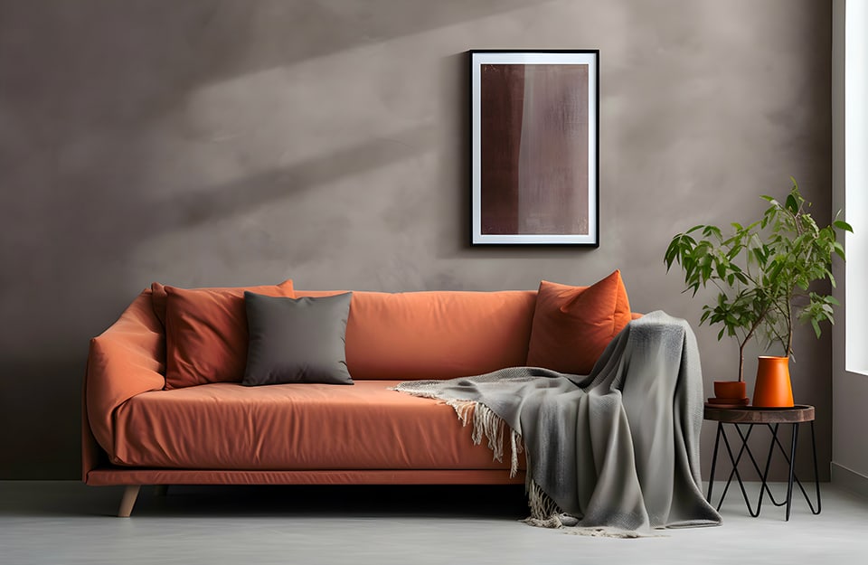 Divano in tessuto color terracotta, con cuscini dello stesso colore e cuscini grigi come la coperta e la parete in cemento della stanza, dove c'è anche un quadro astratto e un tavolino da caffè nero in metallo, con sopra un vaso arancione e una pianta