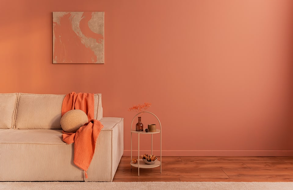 Dettaglio di un salotto con sofà beige, parete color terracotta, richiamato anche da un quadro astratto appeso al muro e da una coperta, e tavolino da caffè minimale in metallo, dello stesso colore del divano, con soprammobili appoggiati
