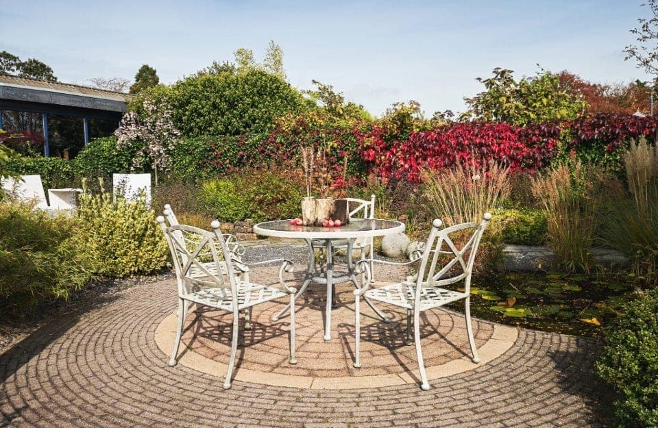 Mesa y sillas de jardín de metal pintado de blanco, sobre un claro con ladrillos de piedra en un jardín con plantas tapizantes y arbustos