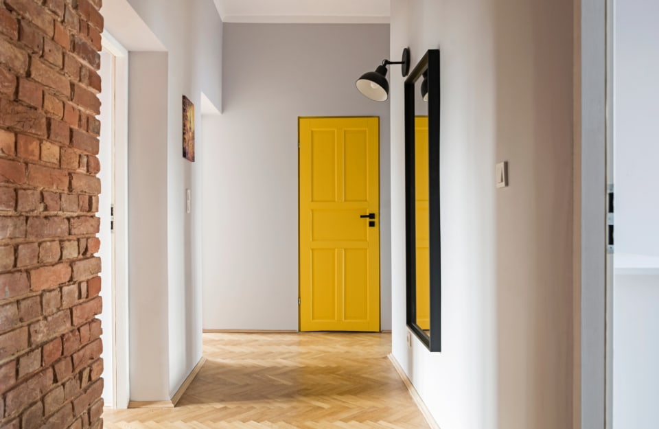A corridor of a house, with several doors facing it and a bright yellow door at the end. The walls are light-coloured, except for one in brick