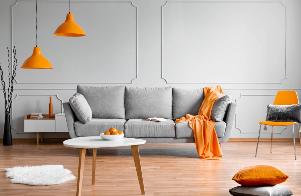 Salotto sui toni del grigio chiaro (pareti, sofà) e con l'arancione zucca come colore d'accento, declinato su lampadari, coperte, cuscini e seduti. Il pavimento è in parquet chiaro