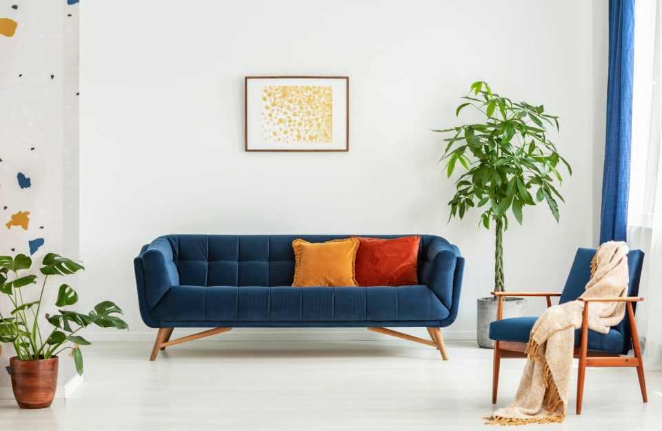 Stanza con parete bianca con divano in tessuto blu in stile modernista Mid-Century con un cuscino giallo e uno rosso, poltrona in legno, quadro astratto sopra il divano e un paio piante