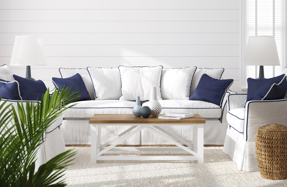 Salotto di una casa in stile coastal, con divani bianchi  con bordature blu navy, cuscini pure bianchi e blu, tavolino da caffè in legno, cestino in vimini, pareti bianche in legno