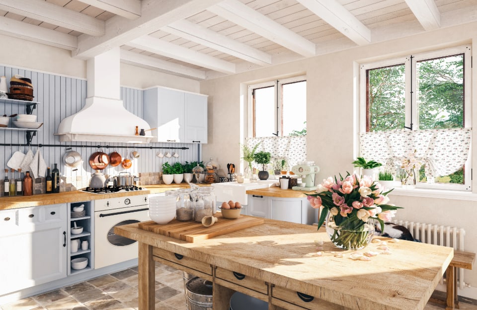 Cucina in stile cottagecore, con tendine rustiche alle finestre, grande tavolo da lavoro in legno, fiori e piante, soffitto con travi in legno bianche