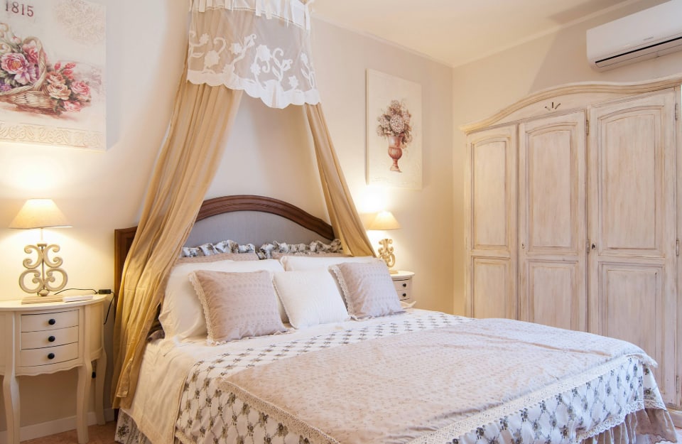 Camera da letto in stile shabby chic, con letto con biancheria romantica, armadio bianco a effetto sbiadito, comodini in stile vintage e quadri florali alle pareti