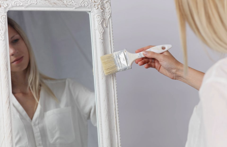 Una donna bionda sta dipingendo in bianco, con un pennello la cornice vintage di uno specchio in cui vediamo anche il suo riflesso