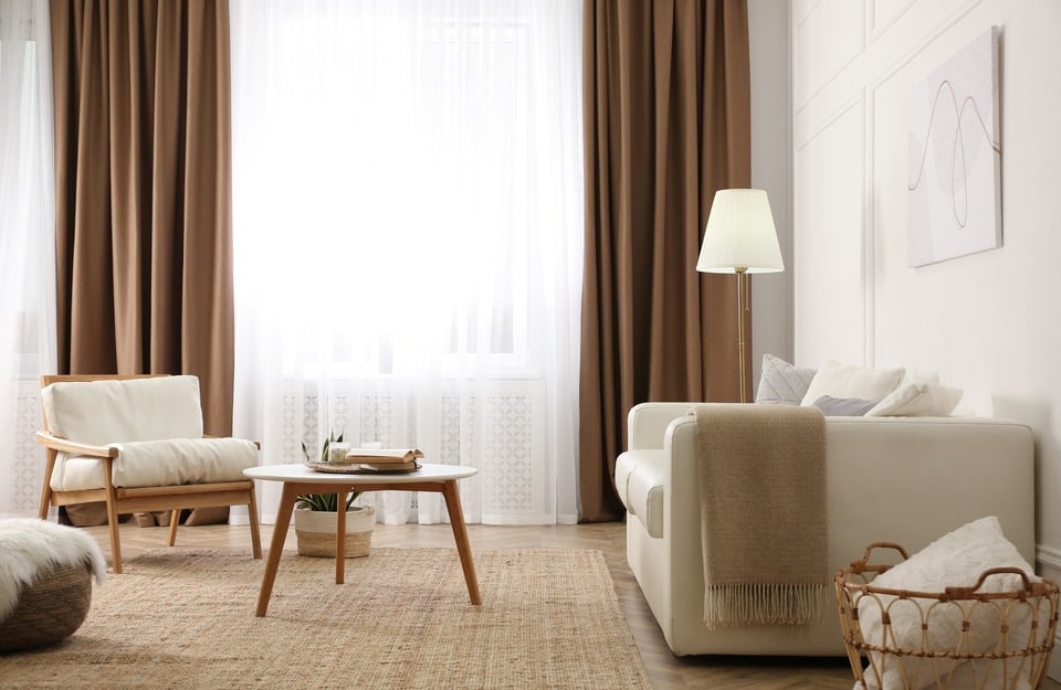 Soggiorno in neutri caldi con tende biscotto, divano chiaro, poltrona in legno e tappeto in fibra naturale. Atmosfera accogliente e luminosa.